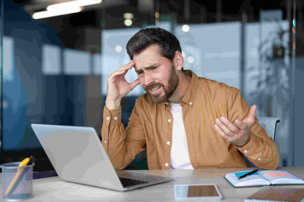 Frustrated businessman in office using laptop with tablet on desk, dealing with stress during work hours