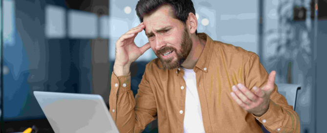 Frustrated businessman in office using laptop with tablet on desk, dealing with stress during work hours